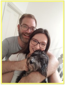 a photograph of a couple, a man and a woman. They are smiling into the camera, both are wearing short-sleeved t-shirts and glasses. The man has his arm around the woman and the woman is holding a black dog that is looking into the camera.
